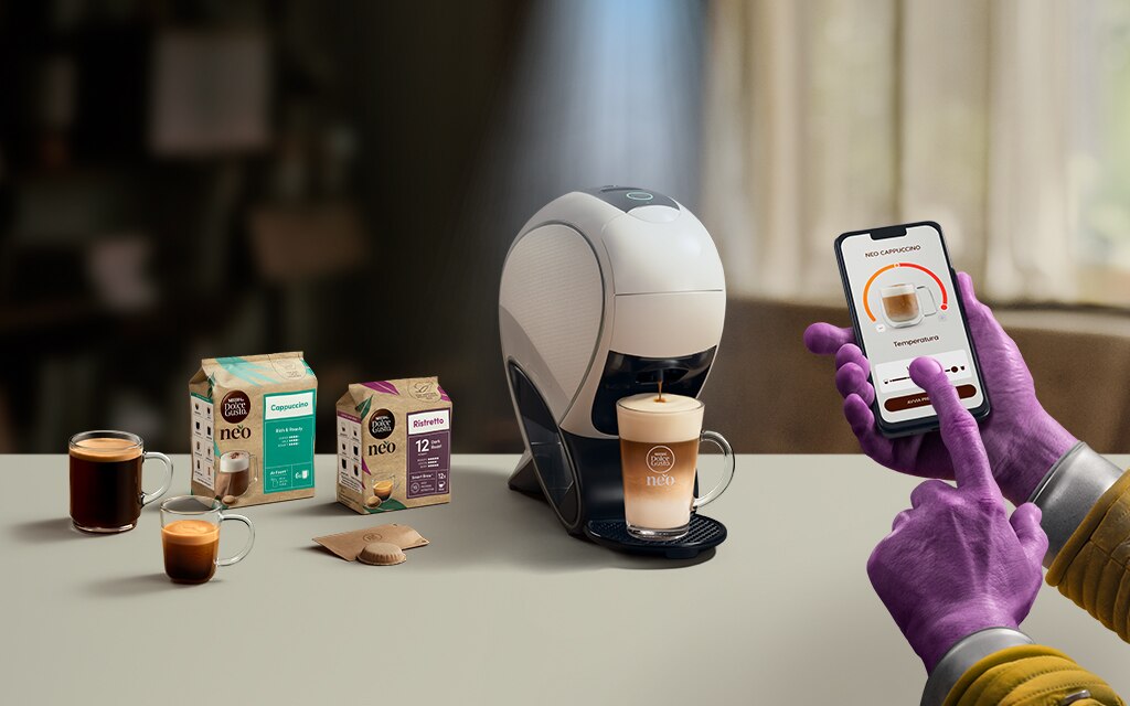 Una mano aliena che usa l'app NESCAFÉ Dolce Gusto per personalizzare un caffè. Macchina da caffè NEO Latte, confezione di caffè e bevande.